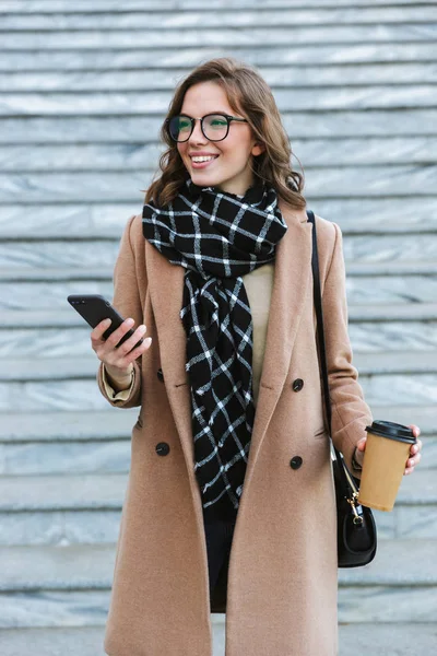 Счастливая молодая женщина прогуливается по улице и пьет кофе с помощью мобильного телефона. . — стоковое фото