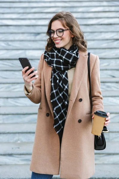 Счастливая молодая женщина прогуливается по улице и пьет кофе с помощью мобильного телефона. . — стоковое фото