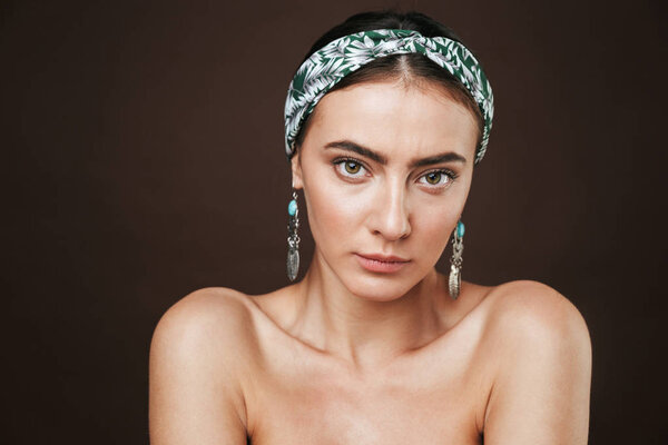 Beauty portrait of a topless young beautiful woman wearing headband and earrings standing isolated over black background