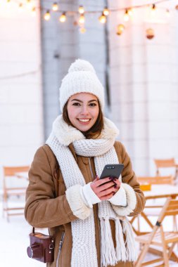 Şapka ve atkı Kışın açık havada yürüyüş genç kadın kar içme kahve istimal hareket eden telefon.