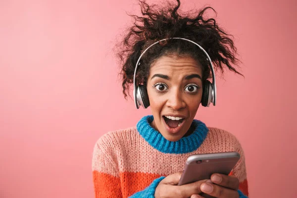 Emocional hermosa joven africana posando aislada sobre fondo de pared rosa escuchando música con auriculares . —  Fotos de Stock