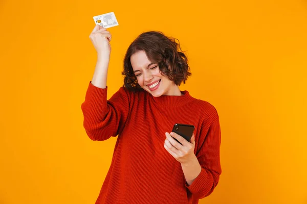 Heyecanlı duygusal genç güzel kadın cep telefonuyla kredi kartı tutan sarı duvar arka plan üzerinde izole poz. — Stok fotoğraf