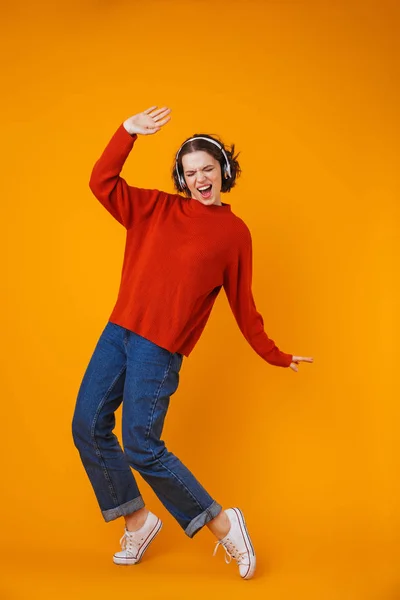 Emocional joven bonita mujer posando aislada sobre fondo amarillo de la pared escuchando música con auriculares . —  Fotos de Stock