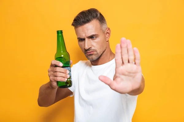 Hombre serio posando aislado sobre fondo de pared amarillo mostrando gesto de stop sosteniendo botella con alcohol . — Foto de Stock