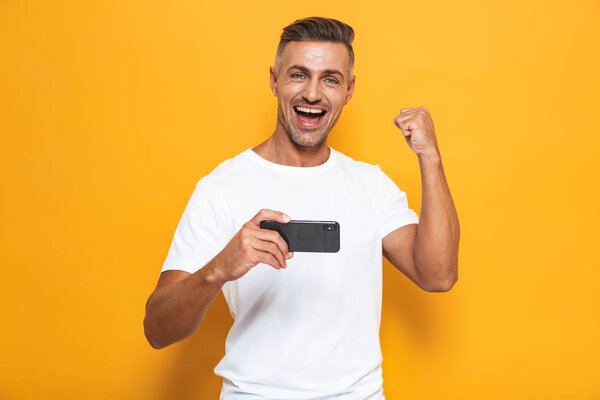 Emotional man posing isolated over yellow wall background play games mobile phone.