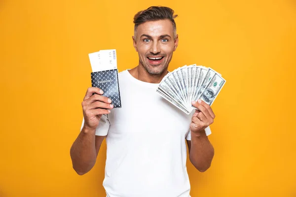 Hombre emocional posando aislado sobre fondo de pared amarilla sosteniendo pasaporte con boletos y dinero . —  Fotos de Stock