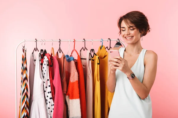 Boldog asszony stylist jelentő elszigetelt felső rózsaszín háttér közelében egy csomó ruhát, mobiltelefon használata. — Stock Fotó