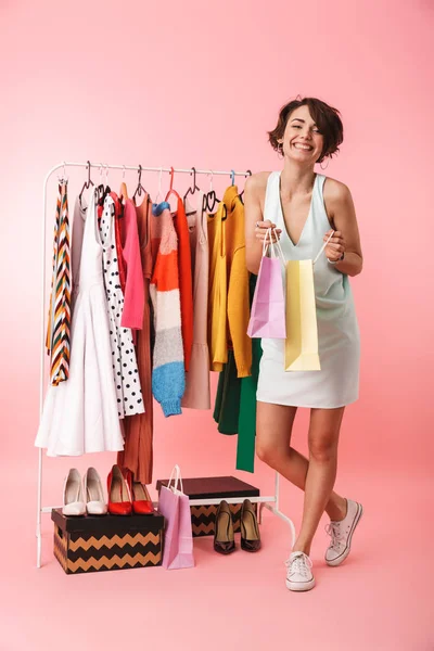 Beautiful woman stylist posing isolated over pink wall background near a lot of clothes holding shopping bags.