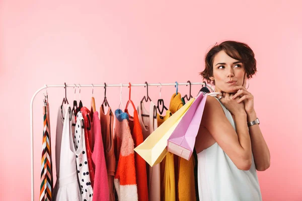 Gyönyörű gondolkodó nő stylist jelentő elszigetelt közelében egy csomó ruhát rózsaszín háttér felett. — Stock Fotó