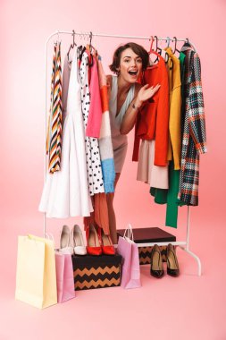 Kadın stilisti, bir sürü kıyafetin yanında pembe duvar arkasında yalnız poz veriyor..