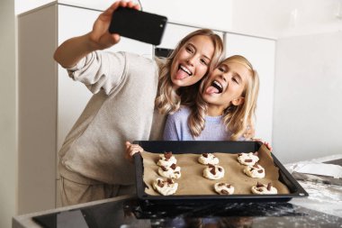 Woman with her little sister indoors at home kitchen cooking sweeties bakery take a photo by phone. clipart