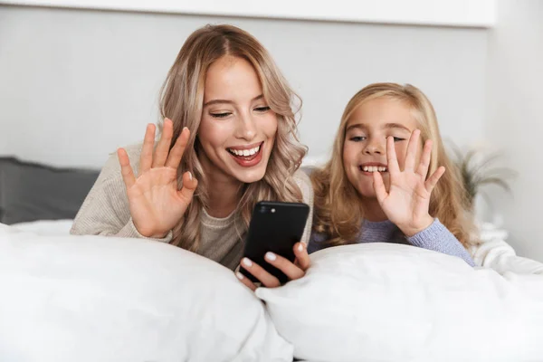 Gelukkig meisjes zusters op bed in de slaapkamer thuis poseren glimlachend zwaaien praten via de mobiele telefoon. — Stockfoto