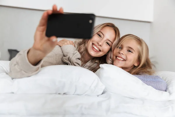 Lányok nővérek hálószobában otthon ágyon pózol, mosolyogva, mobiltelefon használata. — Stock Fotó