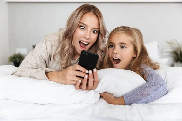 Девушки сестры на кровати в спальне дома позируют улыбаясь с помощью мобильного телефона . — стоковое фото