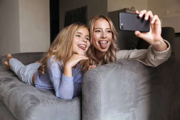 Gelukkig jonge vrouw met haar kleine zusje thuis binnenshuis een selfie nemen per mobiele telefoon. — Stockfoto