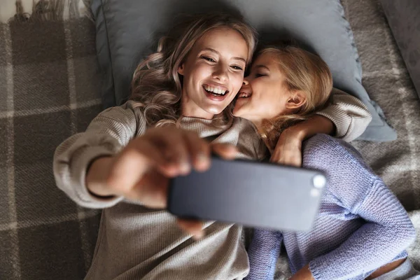 Счастливая молодая женщина со своей младшей сестрой в помещении дома делают селфи по мобильному телефону . — стоковое фото