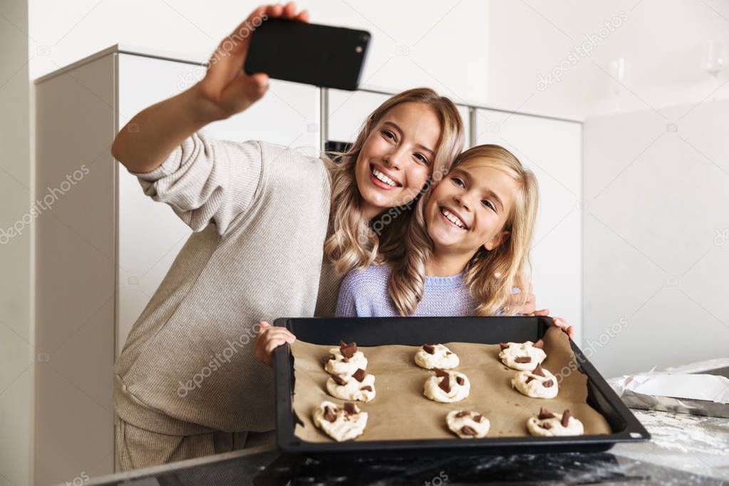 Woman with her little sister indoors at home kitchen cooking sweeties bakery take a photo by phone.