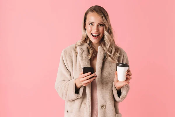 Gelukkig jonge mooie vrouw poseren geïsoleerde over roze muur achtergrond met behulp van de mobiele telefoon koffie drinken. — Stockfoto