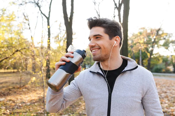 Gefühlsjunger Sportler im Park hört Musik mit Kopfhörern und trinkt Wasser. — Stockfoto