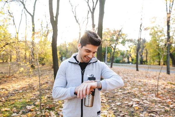 Jeune sportif à l'extérieur dans le parc écoutant de la musique avec écouteurs eau potable regardant regarder . — Photo