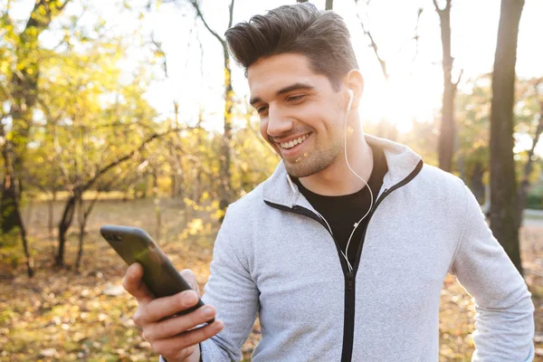 Szczęśliwy młody sportowiec na świeżym powietrzu w parku słuchania muzyki za pomocą słuchawek przy użyciu telefonu komórkowego. — Zdjęcie stockowe