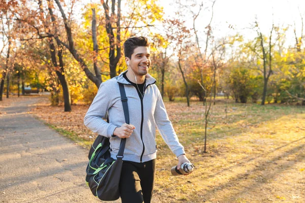 Deportista al aire libre en el parque escuchando música con auriculares caminando con bolsa . —  Fotos de Stock