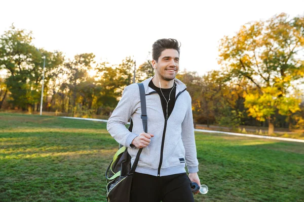 Deportista al aire libre en el parque escuchando música con auriculares caminando con bolsa . —  Fotos de Stock