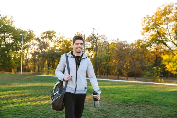 Sportsman outdoors in park listening music with earphones walking with bag.