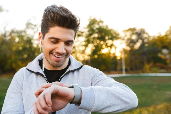 Όμορφος νεαρός αθλητής σε εξωτερικούς χώρους σε πάρκο ακούτε μουσική με ακουστικά, ψάχνουν στο ρολόι. — Φωτογραφία Αρχείου