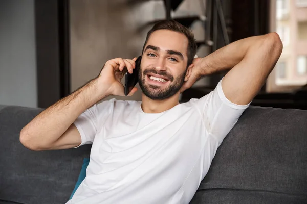 Foto eines brünetten Mannes, der auf dem Smartphone spricht, während er auf sof sitzt — Stockfoto