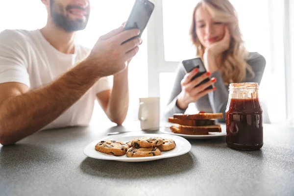 Casal amoroso sentado na cozinha tomar um café da manhã usando telefones celulares . — Fotografia de Stock