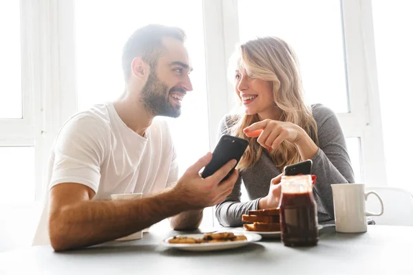Una pareja de enamorados sentados en la cocina desayunan con teléfonos móviles . — Foto de Stock