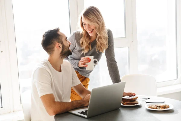 Increíble joven pareja amorosa sentado en la cocina tienen un desayuno con computadora portátil . —  Fotos de Stock