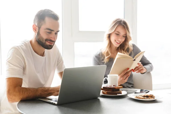 Pasangan luar biasa yang sedang duduk di dapur sarapan menggunakan komputer laptop membaca buku . — Stok Foto