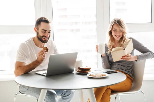 Niesamowite Młoda para kochających siedzi w kuchni zjeść śniadanie przy użyciu komputera przenośnego, czytanie książki. — Zdjęcie stockowe