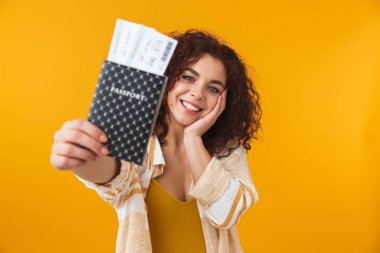 Pasaport ve bilet tutan sarı duvar arka plan üzerinde izole poz sevimli güzel duygusal genç kıvırcık kız.