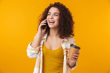 Duygusal genç kıvırcık kız kahve cep telefonuyla konuşurken içme sarı duvar arka plan üzerinde izole poz.