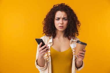 Cep telefonu kullaranarak sarı duvar arka plan içme kahve içerken izole poz memnun kıvırcık kız.