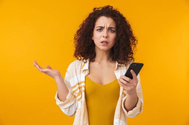 Cep telefonu kullaranarak sarı duvar arka plan üzerinde izole poz karışık memnun kıvırcık kız.