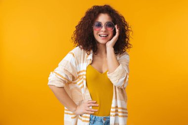 Güzel genç kıvırcık kız güneş gözlüğü takıyor sarı duvar arka plan üzerinde izole poz.