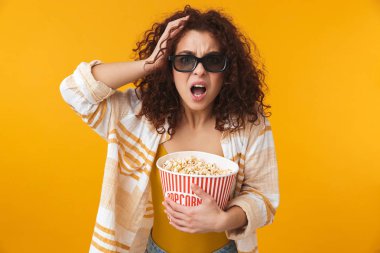 Sevimli güzel genç kıvırcık kız gözlüklü sarı duvar arka plan üzerinde izole poz yemek patlamış mısır film izle.