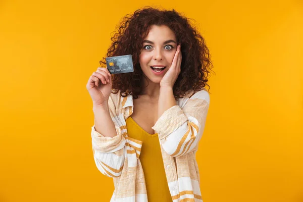 Милая красивая эмоциональная молодая кудрявая девушка позирует изолированно на фоне желтой стены держа кредитную карту . — стоковое фото