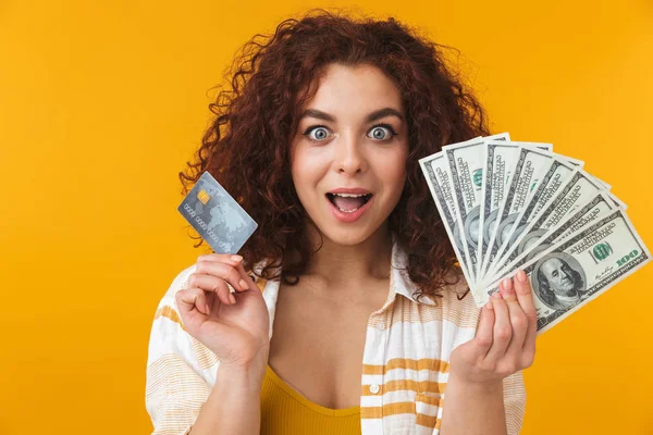 Όμορφη συναισθηματική νεαρή κοπέλα σγουρά θέτοντας απομονωμένες φόντο κίτρινο τοίχο, κρατώντας πιστωτικής κάρτας και τα χρήματα. — Φωτογραφία Αρχείου