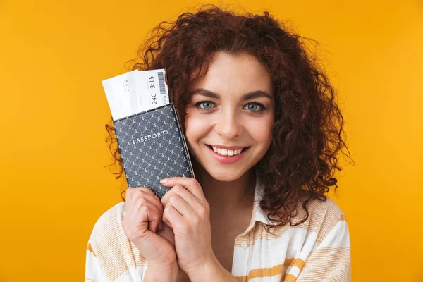 Cute beautiful emotional young curly girl posing isolated over yellow wall background holding passport with tickets. — Stock Photo, Image