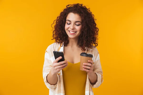Mooie emotionele krullend meisje poseren geïsoleerde over gele muur achtergrond drinken koffie gebruik mobiele telefoon. — Stockfoto
