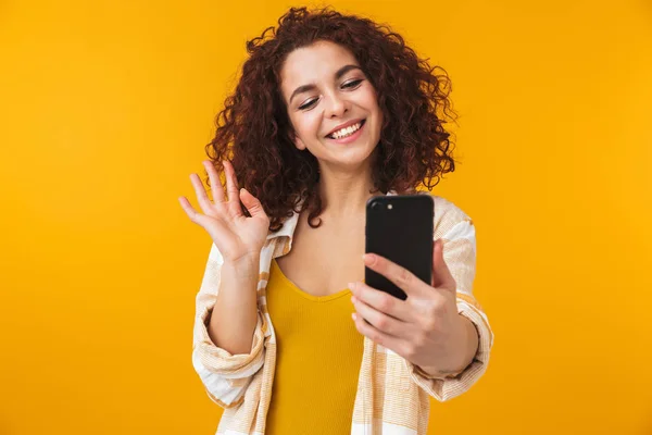 Hermosa chica rizada joven emocional posando aislado sobre fondo amarillo de la pared usando el teléfono móvil . —  Fotos de Stock