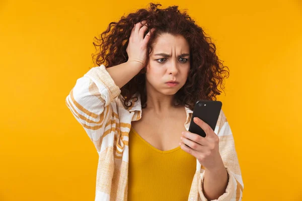Confuso desagradado jovem encaracolado menina posando isolado sobre fundo de parede amarela usando telefone celular . — Fotografia de Stock