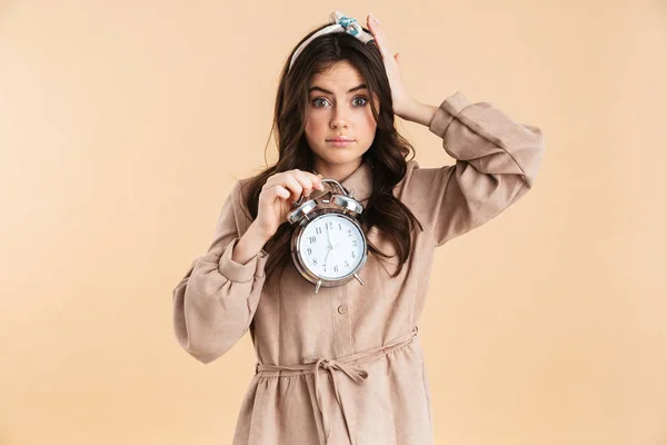 Mujer bonita joven confusa posando aislada sobre una pared de fondo beige sosteniendo el despertador . —  Fotos de Stock