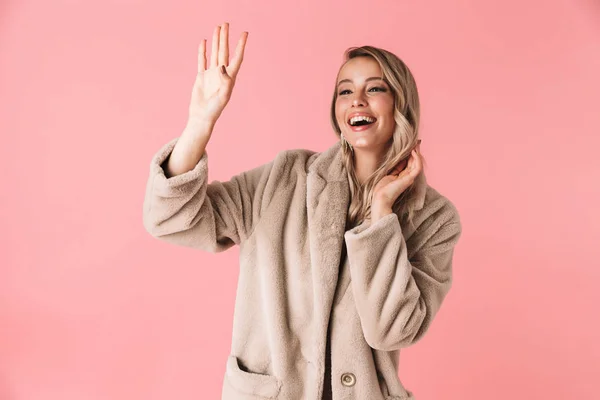 Hübsche Frau posiert isoliert über rosa Wand Hintergrund gekleidet in Modemantel winkt Freund. — Stockfoto