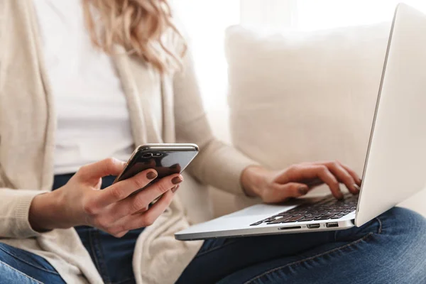 Donna in posa seduta al chiuso a casa utilizzando computer portatile e telefono cellulare . — Foto Stock